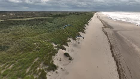 spectacular aerial tracking shot of active paraglider soaring over sandy beach along coastline of sea