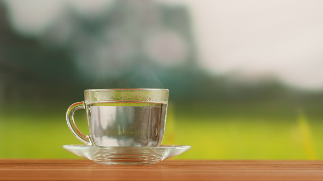 Steaming hot water in a clear glass cup on a wooden table with a blurred green background