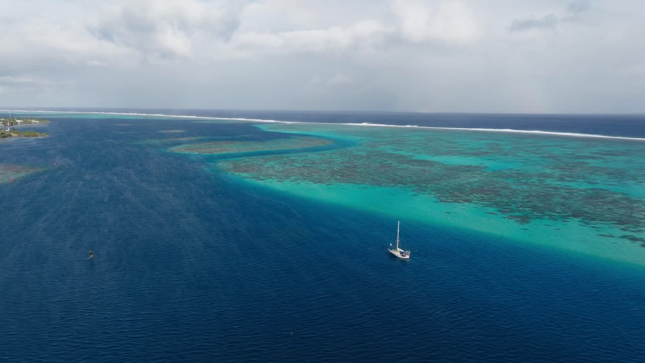 Drone view of a sailboat going through the azure blue waters in Tahiti, French Polynesia. Surrounded by coral reefs on a cloudy day