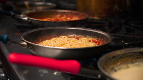 chef stirs tosses and swirls fresh cooked pasta into steaming tomato sauce over italian restaurant prep kitchen stove top, slow motion 4k