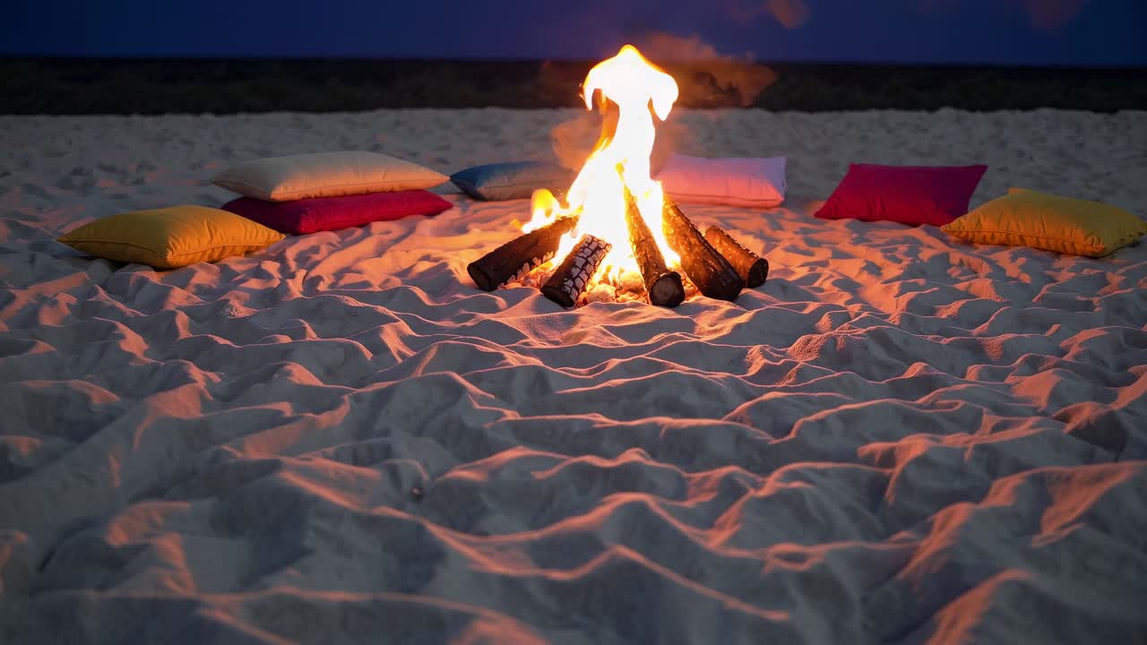 Bonfire burning brightly on a sandy beach at dusk, surrounded by colorful cushions, creating a warm and inviting atmosphere perfect for relaxation and gatherings with friends and family