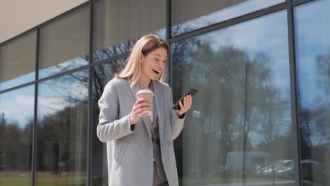 elegant adult happy woman received good news looking at mobile phone screen celebrating successful business deal, high salary. standing near office building. achievement. victory. working results.