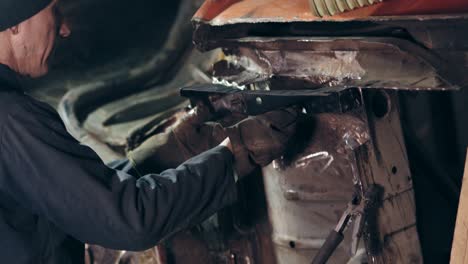 Male-workers-in-workclothes-stand-beside-the-car's-bottom-surface.-One-workman-adjusts-metal-detail-to-car-construction-using