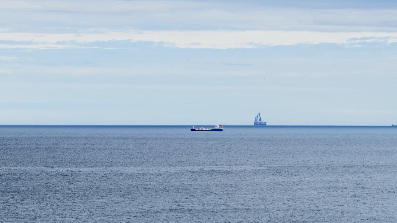 Wide shot of cargo ship passing distant oil rig under overcast sky, tranquil atmosphere
