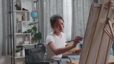 vista lateral de una artista asiática en silla de ruedas sosteniendo un pincel de colores mezclados y pintura en el lienzo