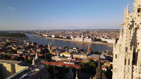 Drone-Volando-Cerca-Del-Campanario-De-La-Iglesia-Matthias,-Revelando-Un-Panorama-Asombroso-De-Edificios-Icónicos-En-La-Orilla-Del-Río-Danubio-En-Plaga