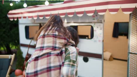 A-happy-brunette-girl-in-a-checkered-shirt-jumps-from-behind-on-her-brunette-boyfriend-in-a-green-checkered-shirt,-and-he-picks-her-up-and-begins-to-spin-her-during-his-vacation-outside-the-city-in-a-camp-near-a-trailer-in-the-summer