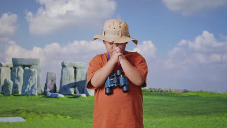 asian boy with a hat and binoculars praying for something while traveling in stonehenge. boy researcher examines something, travel tourism adventure concept