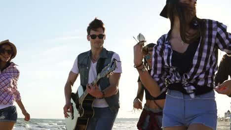 Summer-party-on-the-beach.-Young-boys-and-girls-clapping,-dancing-together-playing-guitar-and-singing-songs-on-a-beach-at-the-water's-edge.-Slowmotion-shot