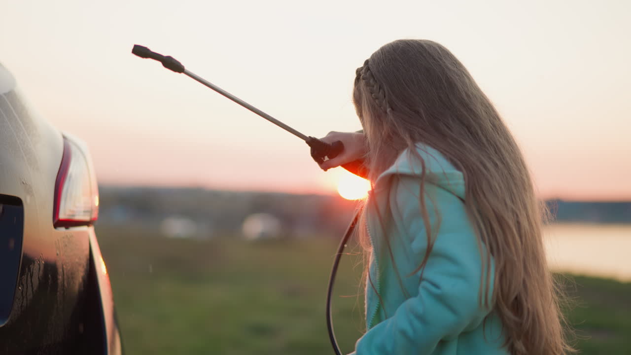 Glatt barn tvättar bil på gräsmark vid solnedgången. Blond liten flicka med spruta torkar droppar från ansiktet.