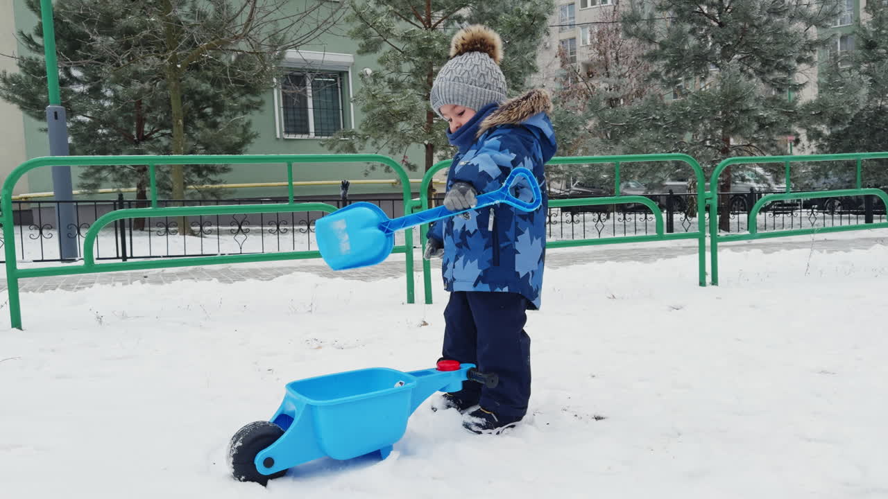 Caucasian toddler in warm winter suit stands outdoors. Lovely male kid picks up snow with his shovel.