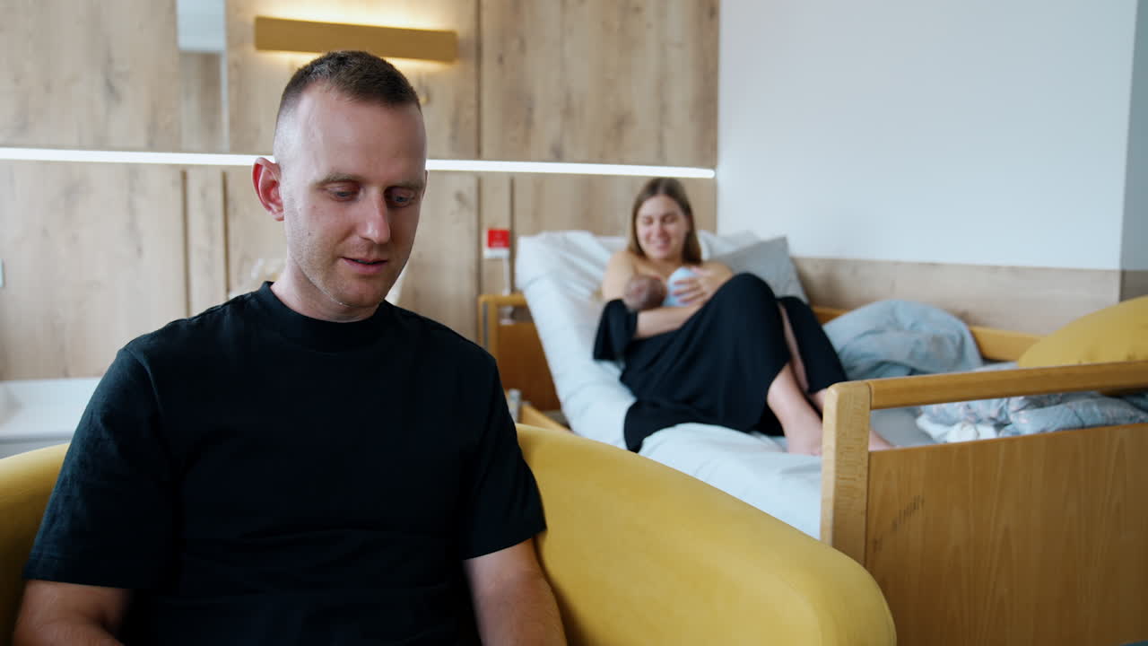 People are in the modern hospital ward after childbirth. Woman holding a baby sitting on the bed and man works at laptop.