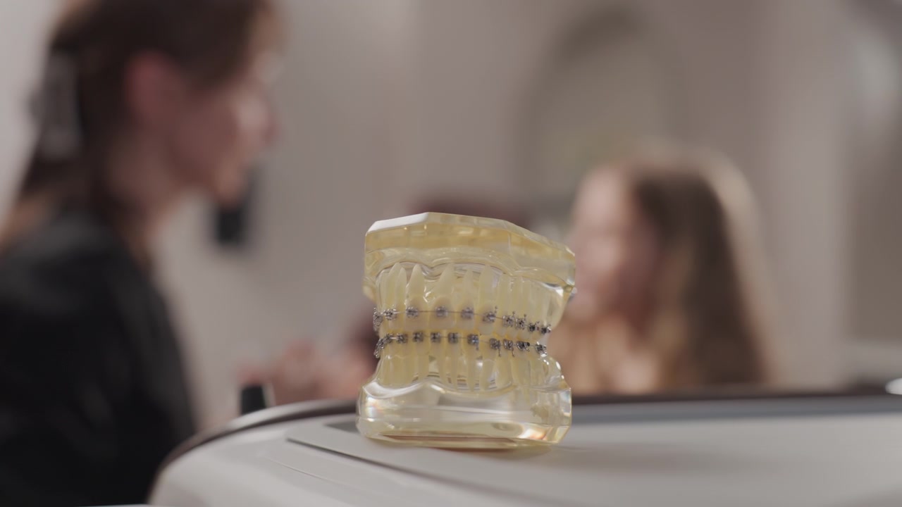 Primo piano di un modello dentale in plastica trasparente con apparecchio ortodontico in mostra in una clinica ortodontica. Una donna e una bambina sono visibili fuori fuoco vicino a uno specchio sullo sfondo. La scena è illuminata delicatamente.