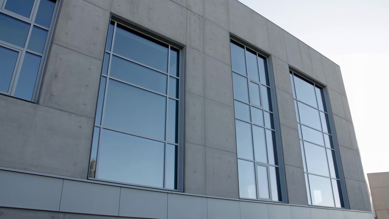 Vista esterna dal basso di un moderno edificio commerciale con tre alte vetrate rettangolari incassate in pannelli di cemento prefabbricato liscio, che mostrano linee pulite, simmetria e texture urbana.