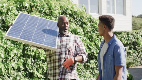 feliz padre afroamericano con panel solar hablando con su hijo adulto en un jardín soleado, cámara lenta