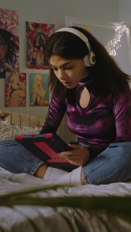 beautiful african american girl in headphones sits on bed in her room and surfs internet using tablet. happy teenager chats with friends or listens music. vertical shot