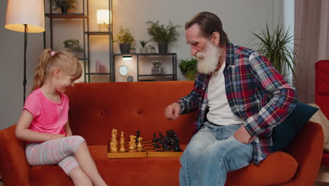 Smiling-senior-grandfather-playing-chess-board-game-with-teen-granddaughter-child-girl-kid-at-home