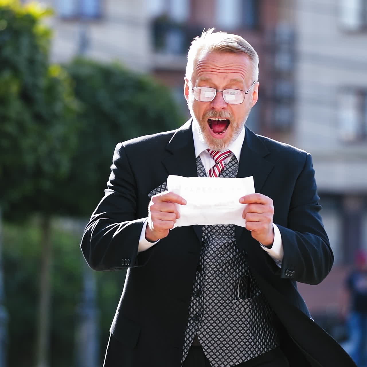 Close-up portrait of a senior man reading paper outdoors. Mature businessman with beard and glasses becomes happy after reading successful information. Slow motion.
