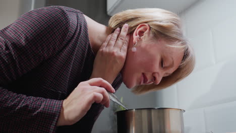 lady with short blonde hair cooking in modern kitchen, bending down to smell food aroma with satisfied smile, hand on neck adorned with pink beaded bracelet, holding ladle over steaming pot