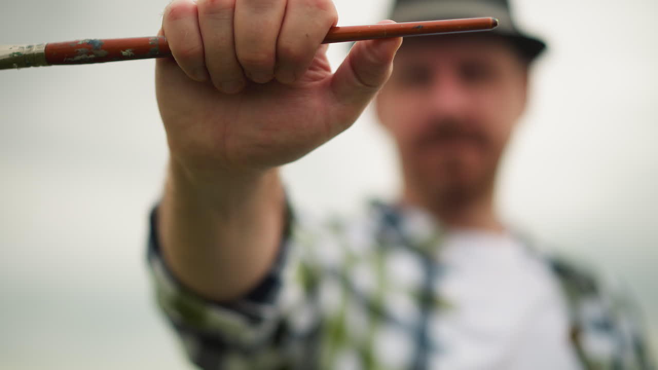 Extreme close-up of a painter's hand gripping a small brush, turning it slightly, with the artist and background completely blurred for focus on the brush