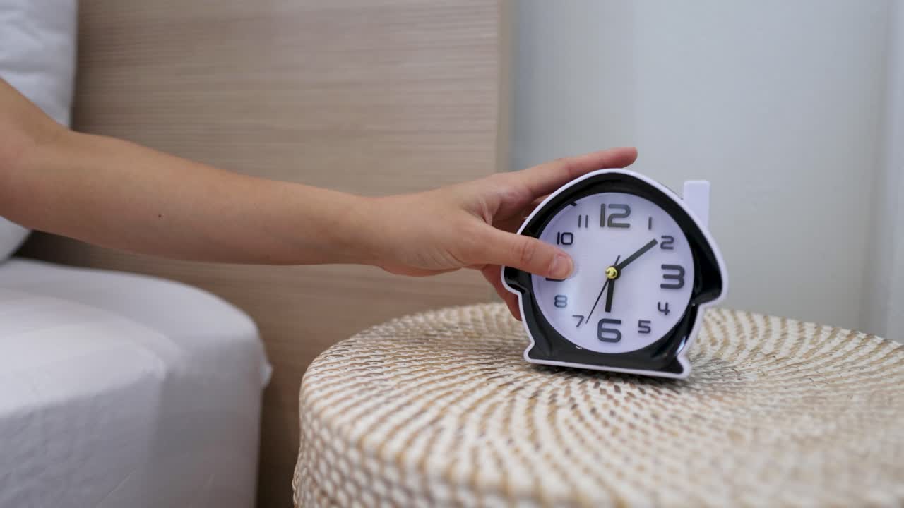 Una mano posa una sveglia nera su un comodino intrecciato in una camera da letto luminosa
