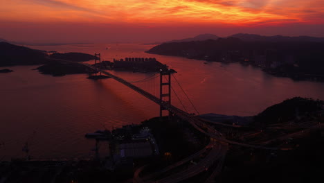 cinematic aerial view of tsing ma bridge in hong kong at sunset, spanning over calm waters with city skyline, mountains, and ships in the distance, creating a serene urban and natural landscape scene