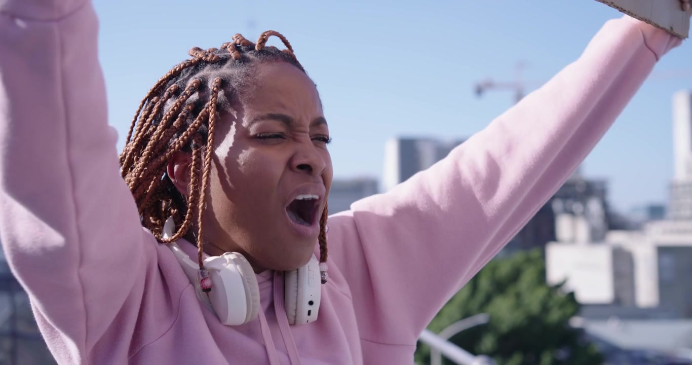 African woman in pink hoodie headphones raising cardboard sign, chanting, demanding change on roof