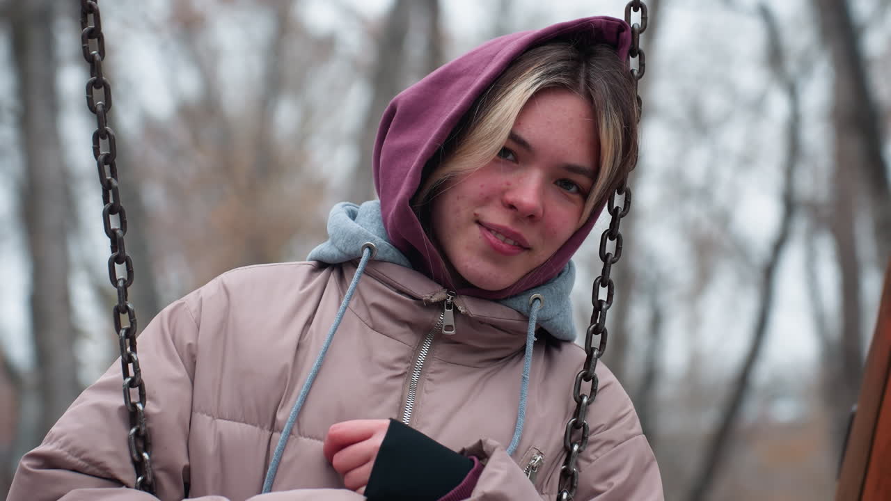 joven con traje de invierno con manchas en la cara sosteniendo una cadena de columpio y apoyando su cabeza en ella, con una chaqueta con capucha, expresión relajada, fondo nevado borroso con árboles desnudos