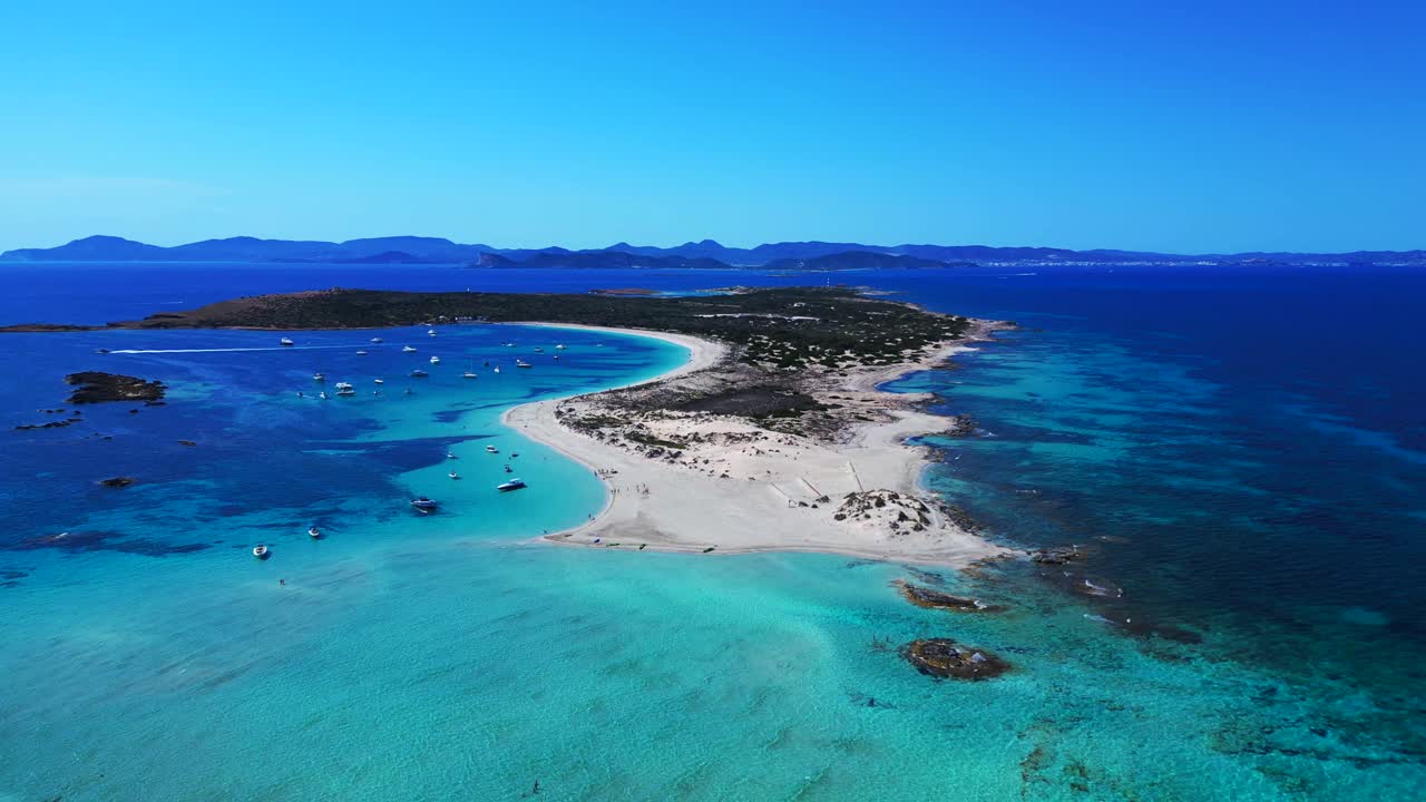 Espalmador island featuring white sand beaches, clear turquoise water, and anchored boats. Ibiza and the island of Es Vedra in the background. Great aerial view flight panorama orbit drone