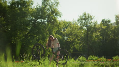 young female cyclist rotates front and back tires of upside-down bike while rotate the pedal too beside lush garden, as blurred pedestrian strolls in background