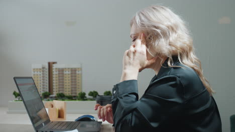 side view of professional lady retrieving phone from folder, placing call while browsing property listings on laptop at organized desk with real estate building models in background