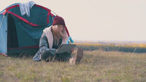 girl hiker in hat with tablet sits at blue tent in evening