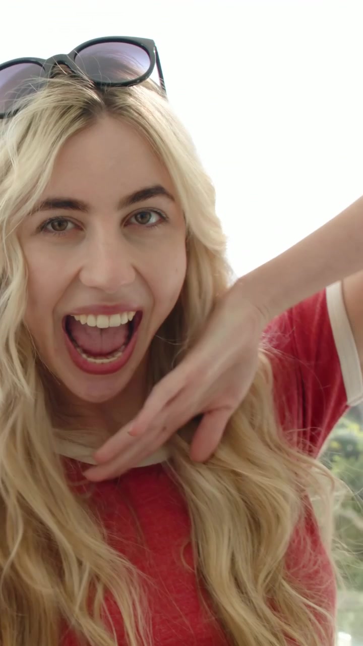 Vertical video: Smiling woman posing on balcony by glass rail as camera pans, in red tee and shades