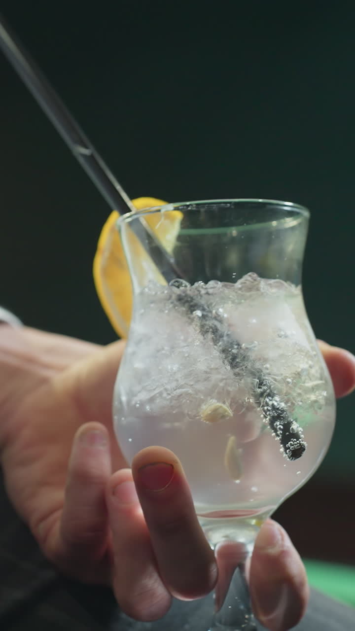 Primo piano di un uomo, volto non visibile, che indossa una camicia bianca e pantaloni grigi, mentre tiene in mano una stecca da biliardo e un bicchiere di limonata con cannuccia nera. Cubetti di ghiaccio e fetta di limone aggiungono dettagli rinfrescanti.