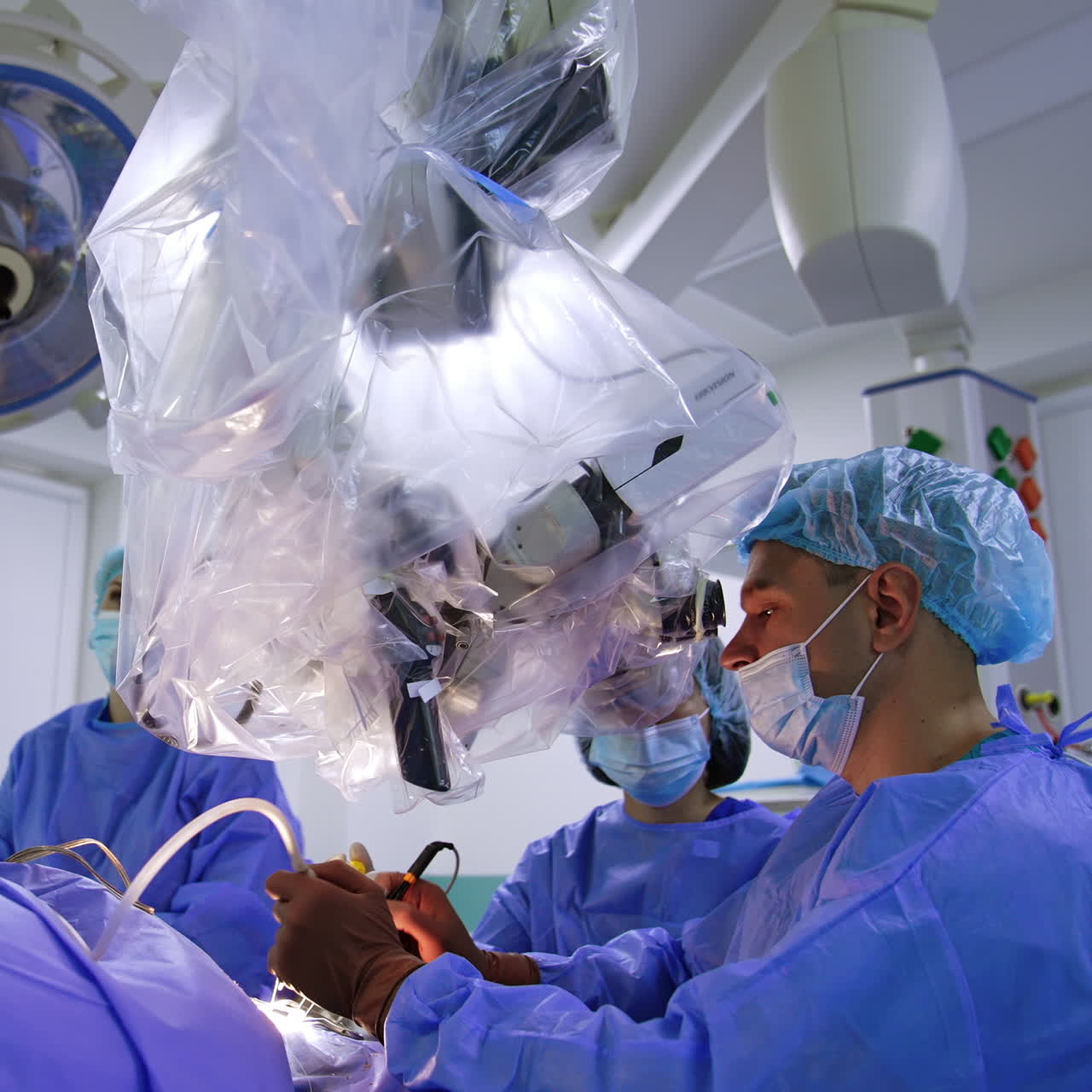 Male neurosurgeon and assisting female doctor look at microscope operating patient. Brain MRI scans on the screen at backdrop.