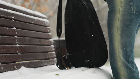 partial view of lady removing guitar pack to place on snow-covered bench in snowy park, with blurred figures walking in distant background surrounded by frosted trees