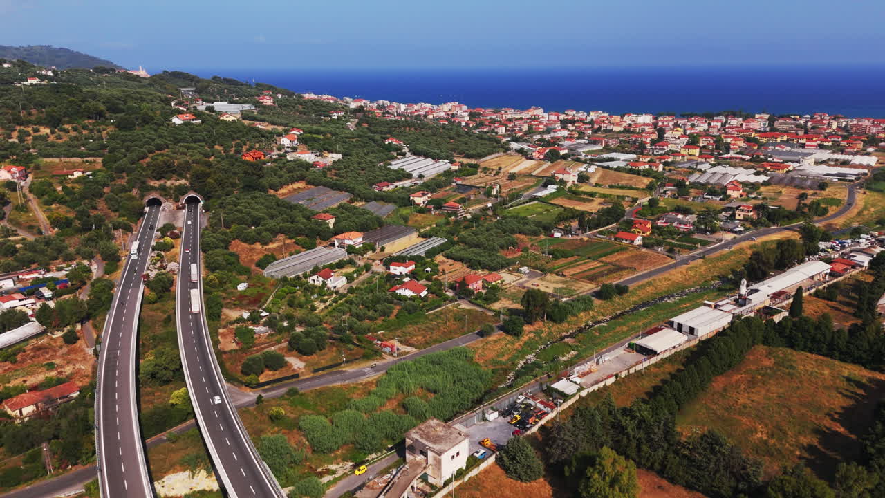 Drone flies forward over highway bridges and tunnels, approaching the coastal city with sea in the background and farmlands below