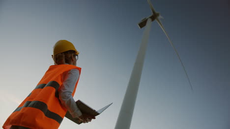 Lower-view-of-a-caucasian-female-engineer-wearing-a-helmet-and-using-laptop-under-the-huge-windmill-turbine-and-watching-it-working-and-producing-energy