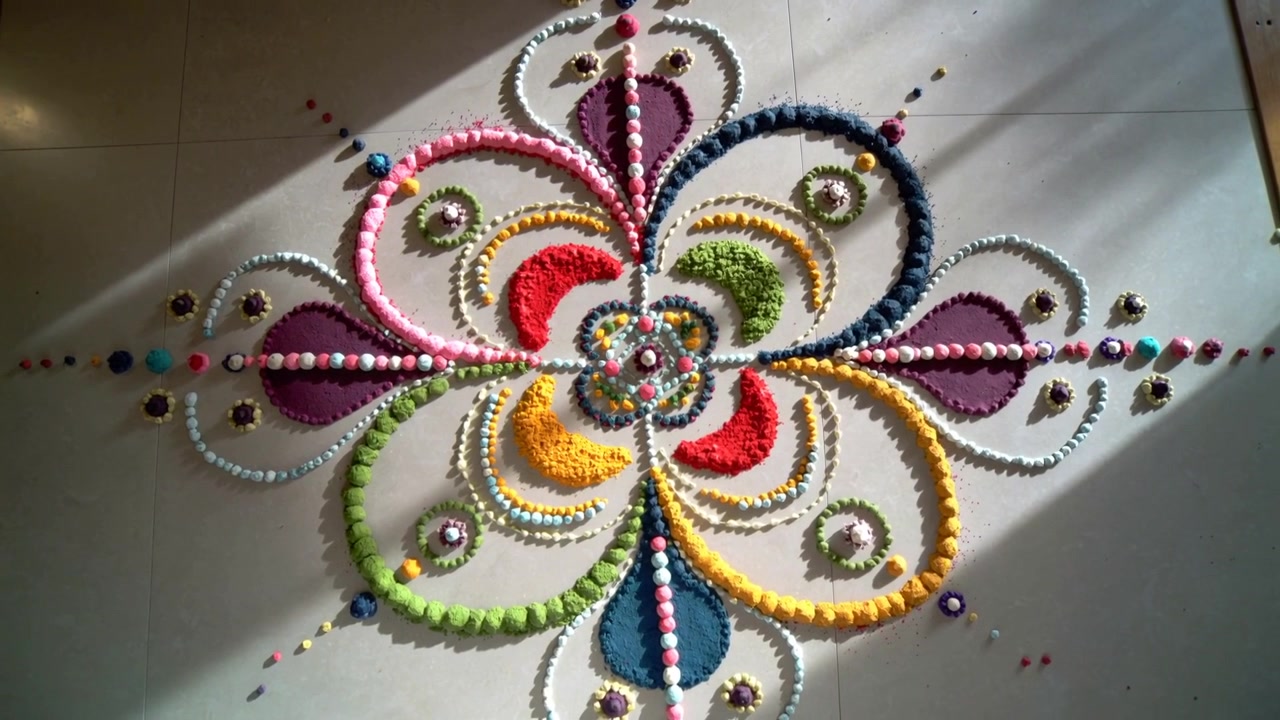 Telecamera che si inclina rivelando un rangoli ornato e multicolore su un pavimento piastrellato, con la luce del sole che si sposta sulle perline