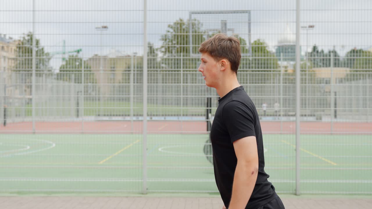 A young man performs squats outdoors, with a football field visible in the background