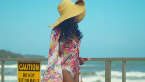 a young black girl in a bikini walks and looks at the ocean in the distance as the waves crash on a sea wall