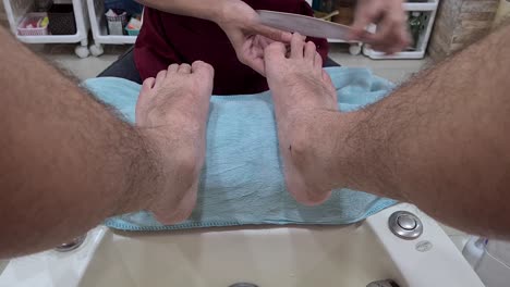 a pov shot of a man receiving a pedicure on his toes, from a woman wearing a red overall