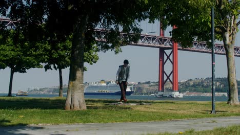 personas relajándose en un parque junto a un río con un crucero y un puente en el fondo