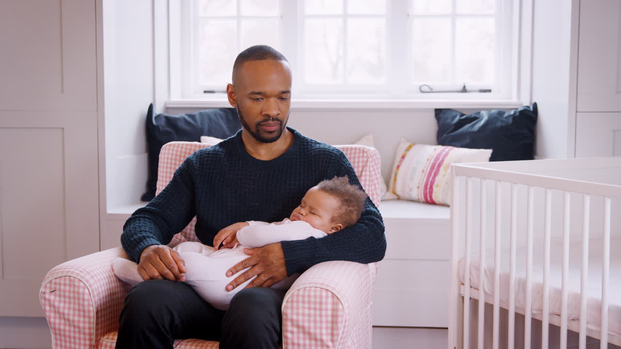 Stressed New Father Sitting In Chair Holding Sleeping Baby Girl In Nursery At Home