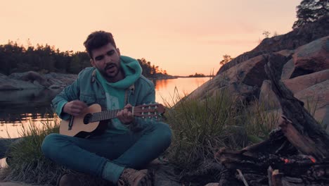 young musician playing guitar and singing in front of bonfire at the ocean. guy is singing in front of friends during sunset while traveling.