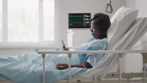 Paciente-Mujer-Negra-En-El-Hospital-Con-Bomba-De-Infusión-Volumétrica-De-Solución-Salina-Usando-Un-Teléfono-Móvil-En-La-Camilla-De-Examen.-Mujeres-Africanas-Acostadas-En-La-Cama-De-Un-Hospital-Con-Un-Teléfono-Móvil-Inteligente-Mientras-Están-En-El-Hospital.