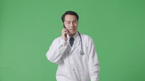 아시아 남성 의사가 스테토스코프를 들고 병원에서 녹색 화면 배경에서 는 동안 스마트폰으로 이야기하고 있습니다.