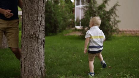 Father-and-son-play-hide-and-seek-around-tree.-Little-blonde-boy-is-running.-Happy-time.-Parenthood.-Green-grass-around