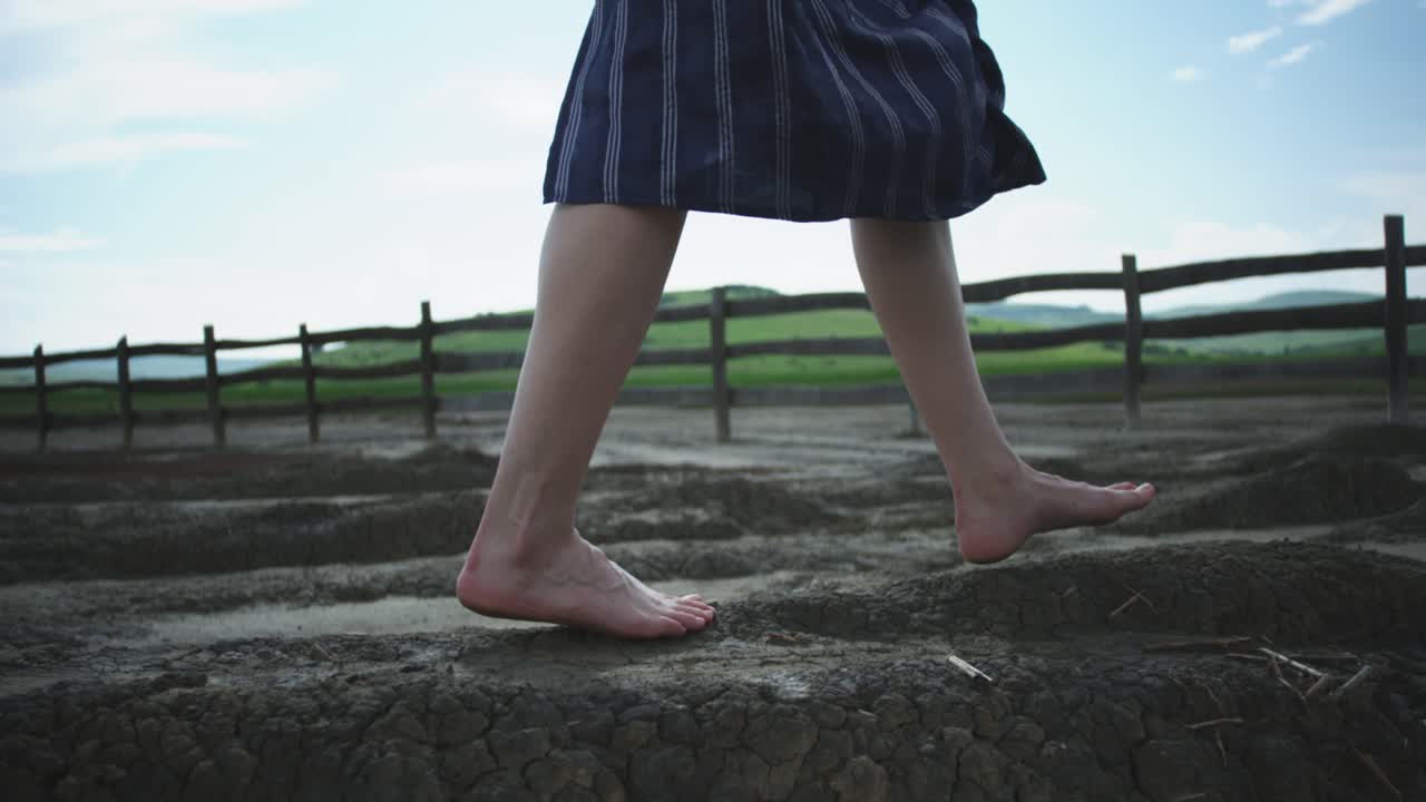 A low-angle close-up follows a woman's bare feet as she walks across the dry, and cracked earth of a mud volcano. This shot captures a sense of freedom and sensory connection with a strange landscape.