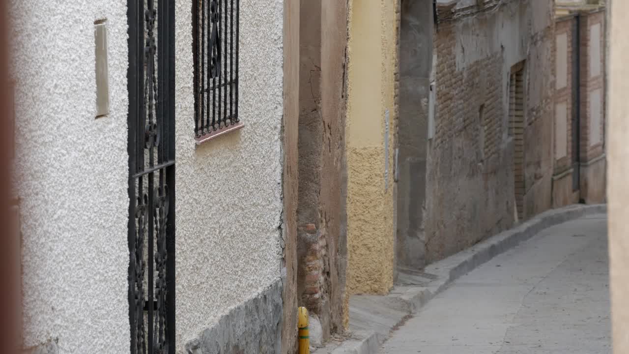 Handheld medium black cow from running on street at traditional celebration, encierro, in Spain. Village one-storey town houses, visible brick on walls. Metal gate on doors, windows.